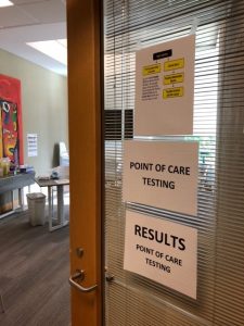 image of the door to the point of care testing room at the Dr peter centre - Signs read Point of Care testing and Results - point of care testing on sheets of white paper.