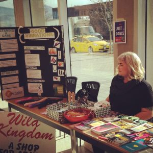 Kate, with blond hair, sits at a table of harm reduction supplies with a three-fold poster board standing next to it.