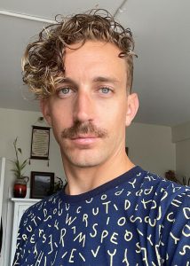 Headshot of Joel who has curly brown hair to the right side of his face, a moustache, and is wearing a t-shirt with scattered alphabet letters.
